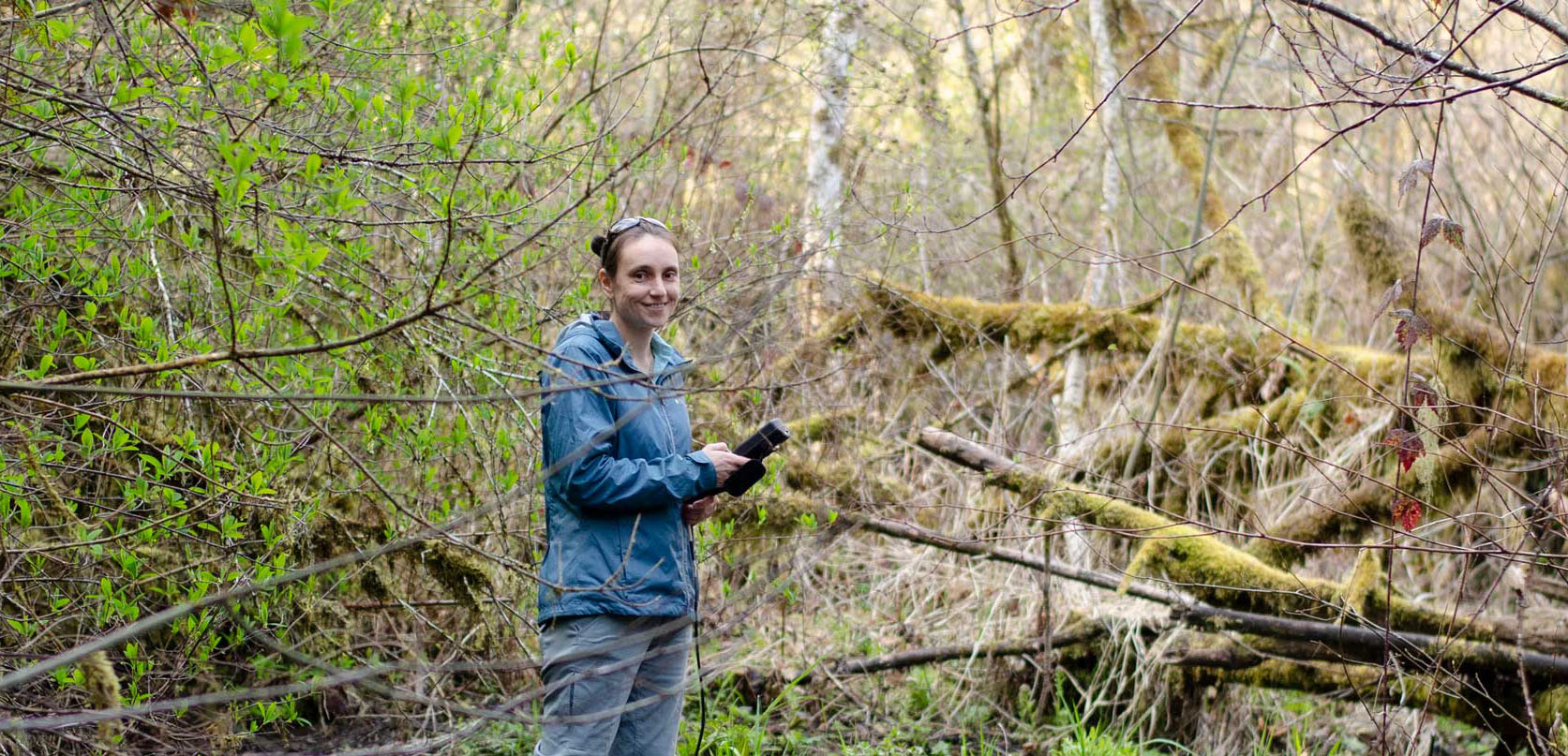 A woman stands in a lush, mossy Oregon forest with a GPS device, surveying the landscape during a woodland field study.