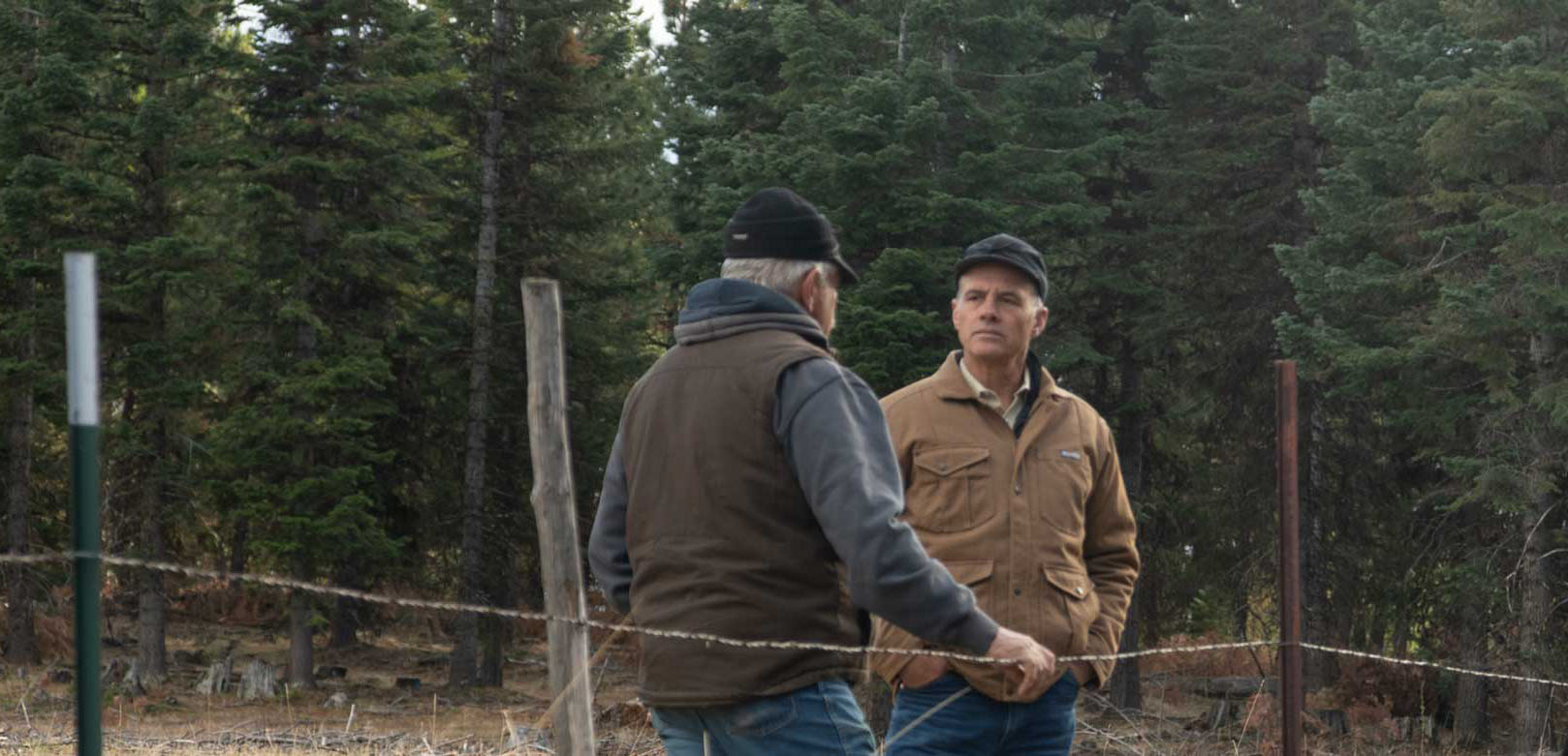 Two men in jackets and hats stand in a forested area having a discussion near a barbed-wire fence.
