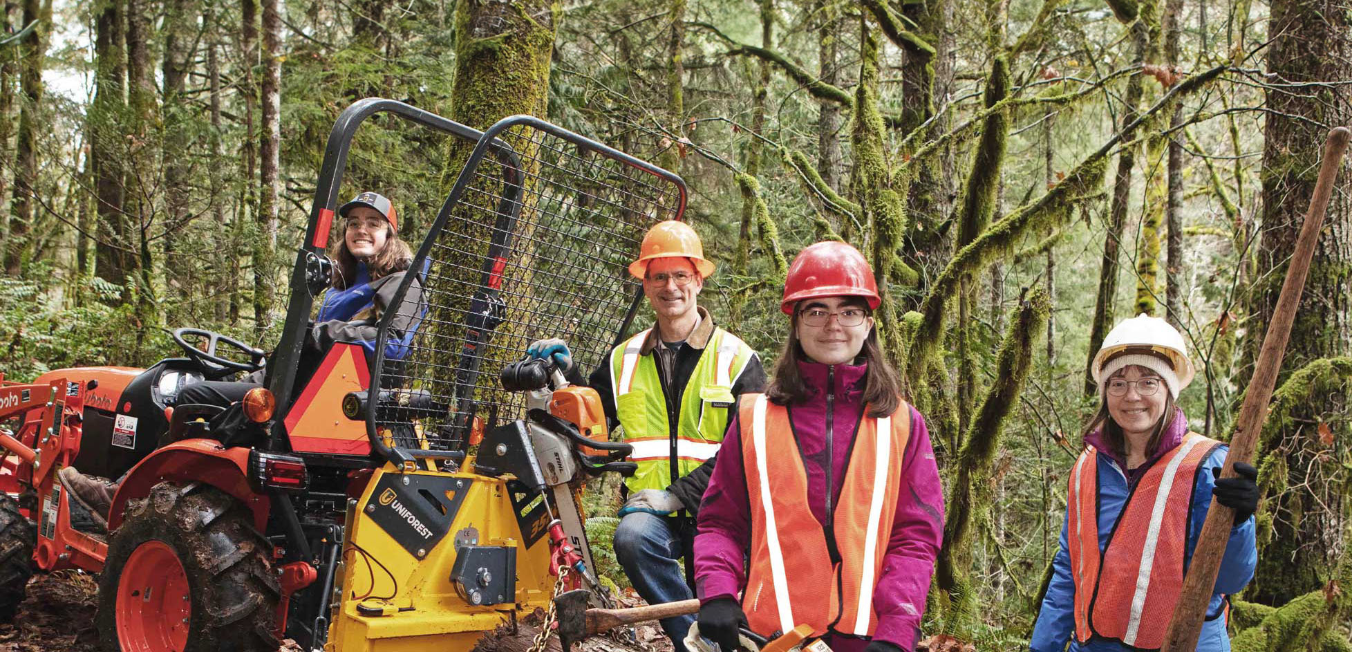 Four people in safety gear, including hard hats and reflective vests, pose in a forest next to a small logging tractor and equipment.