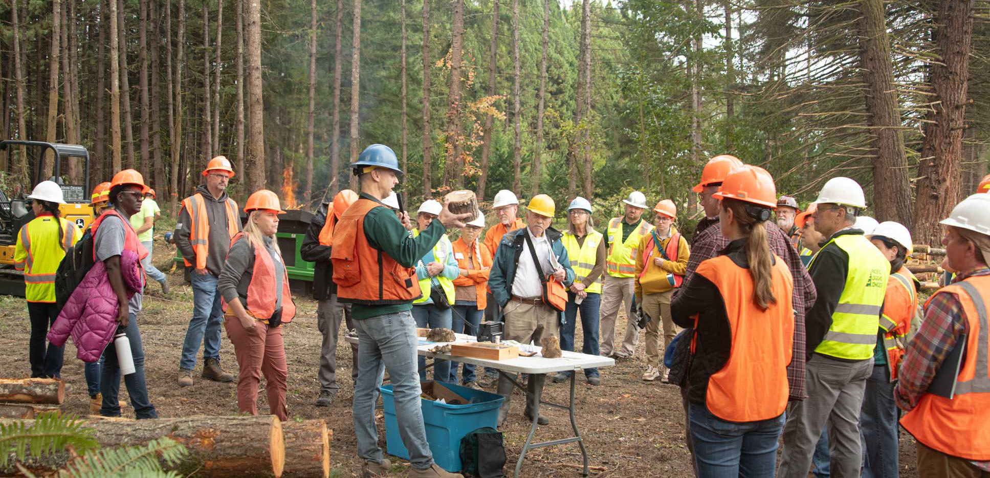 A forestry instructor demonstrates wood characteristics to attendees wearing safety gear at a landowner debris burn event in a forest setting.