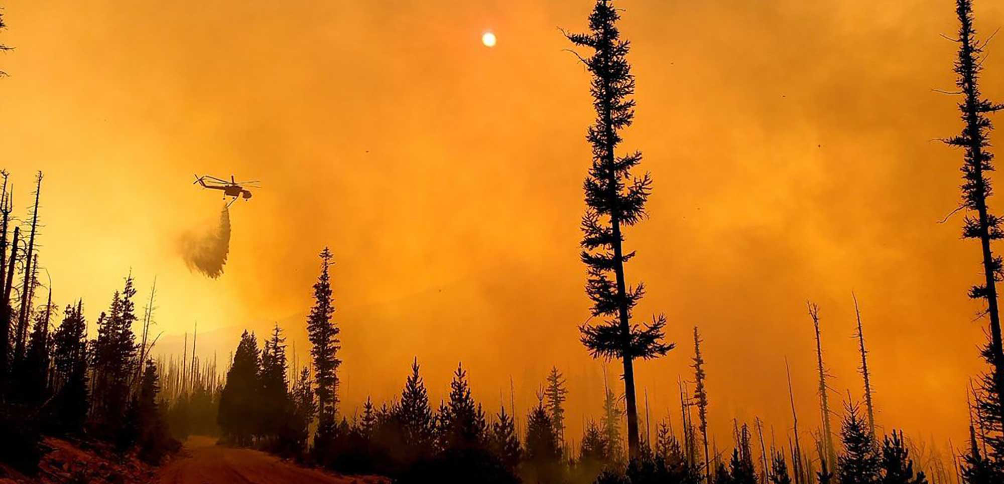 A helicopter drops water over a smoky forest fire under an orange, haze-filled sky.