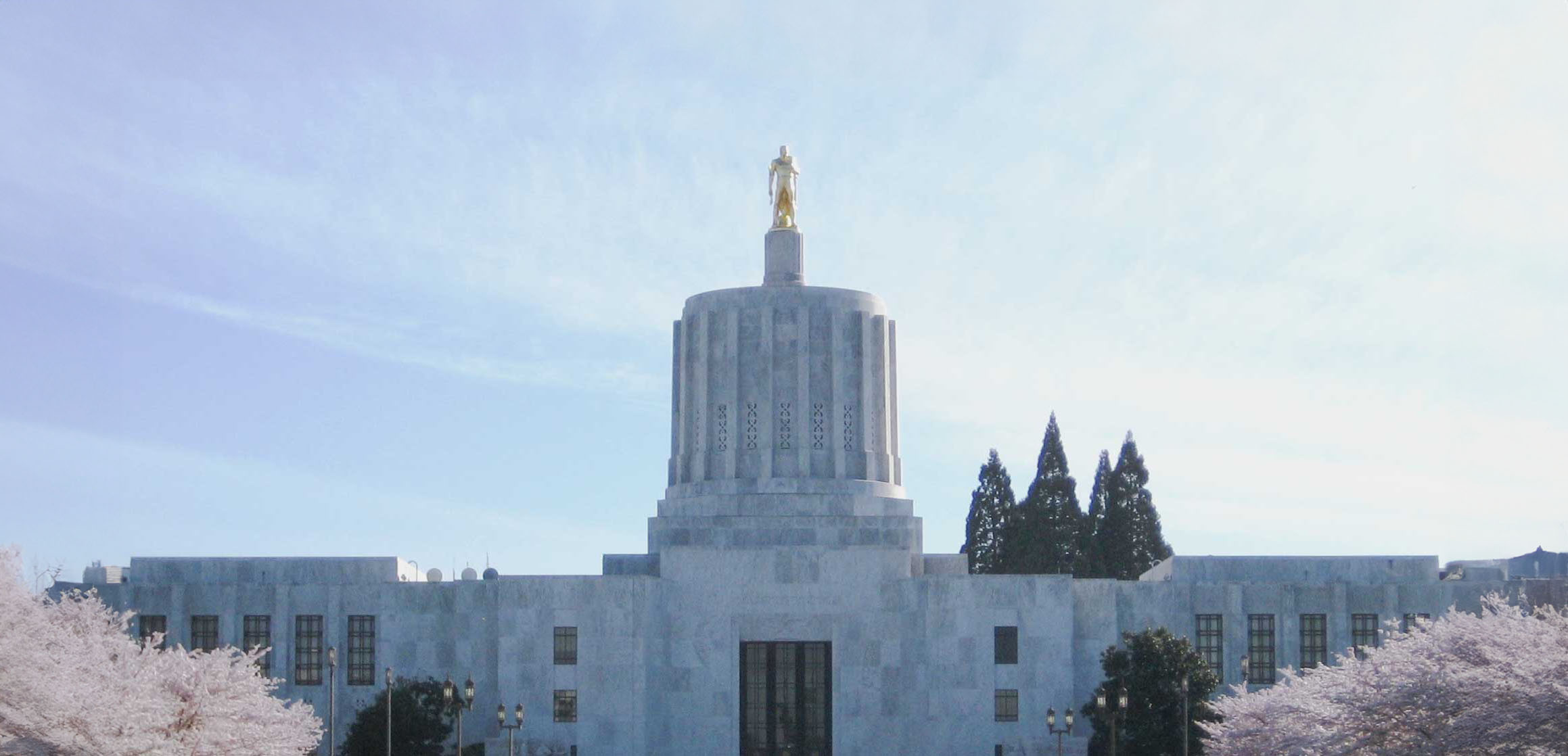 The Oregon State Capitol building in Salem stands beneath a blue sky, crowned with the gold Oregon Pioneer statue.