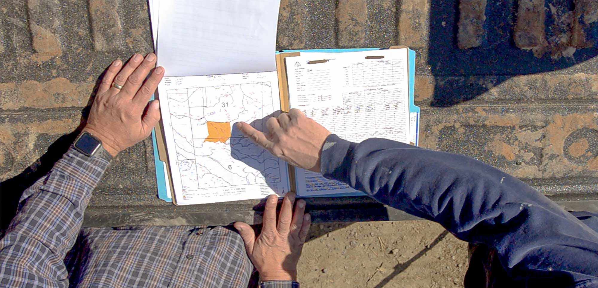 Two people reviewing maps and documents on a truck tailgate, with one person pointing to a highlighted area on the map.
