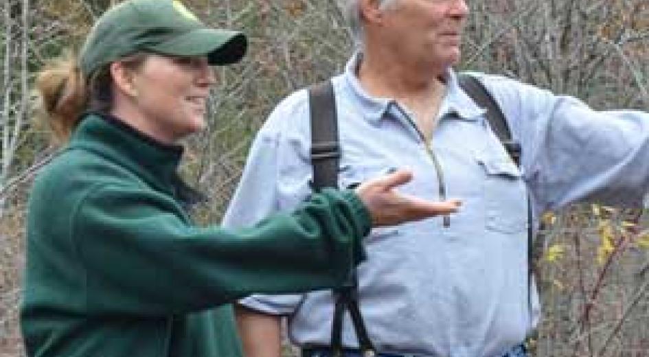 A woman in a green jacket and a man in suspenders have a discussion while standing in a wooded area.