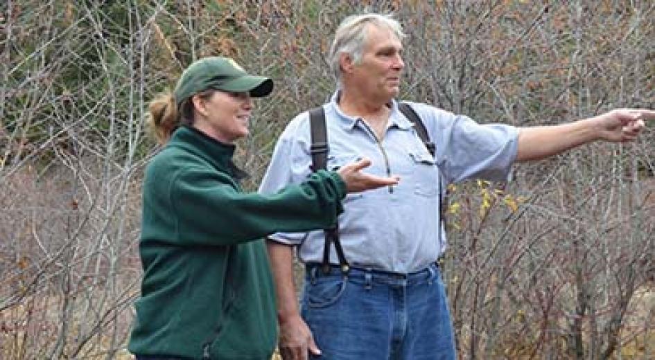 A woman in a green jacket and a man in suspenders have a discussion while standing in a wooded area.